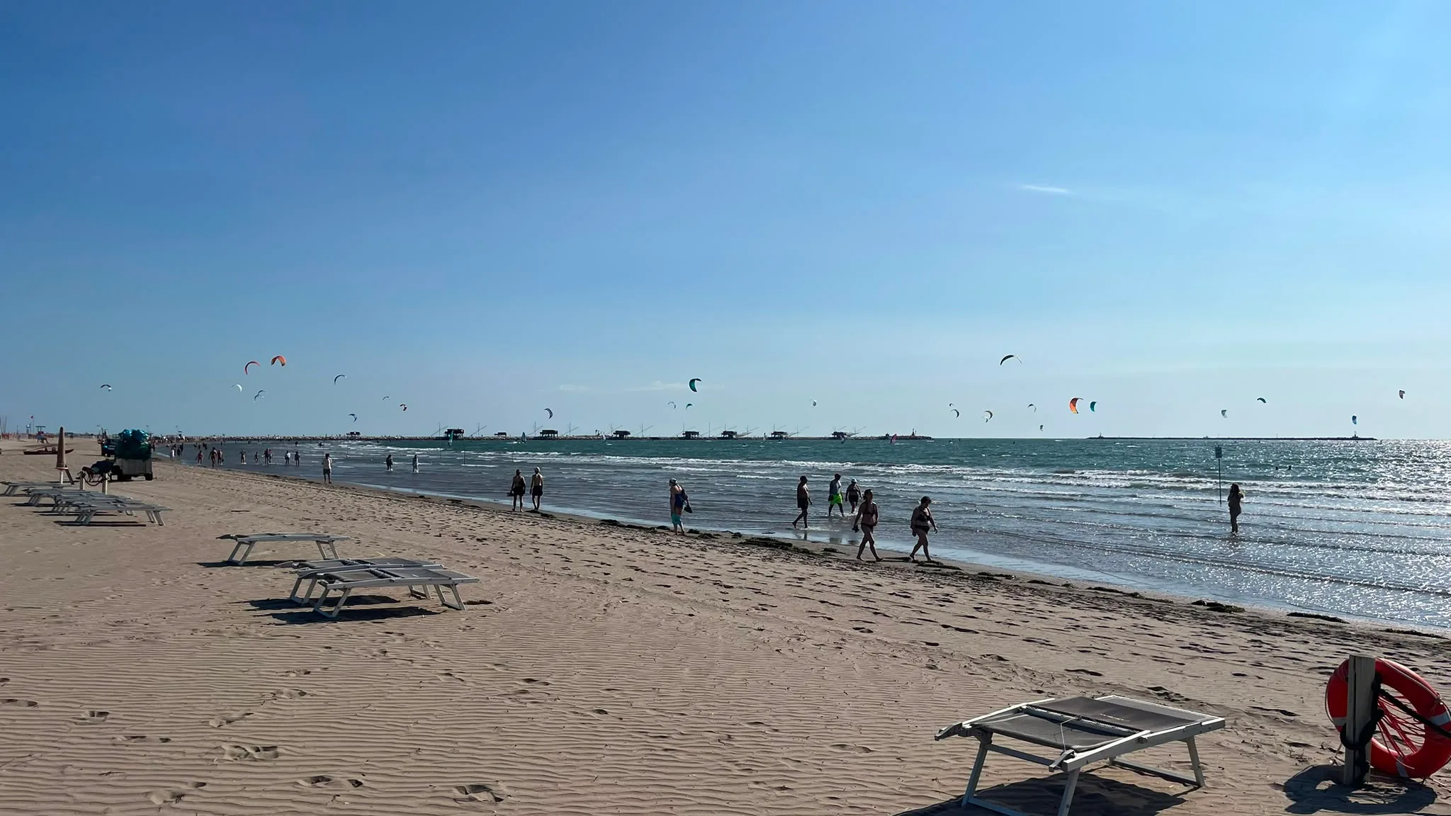 Spiaggia con lettini, corda e boa di salvataggio in primo piano, persone sulla riva e kite surfisti in mare sotto la luce del sole