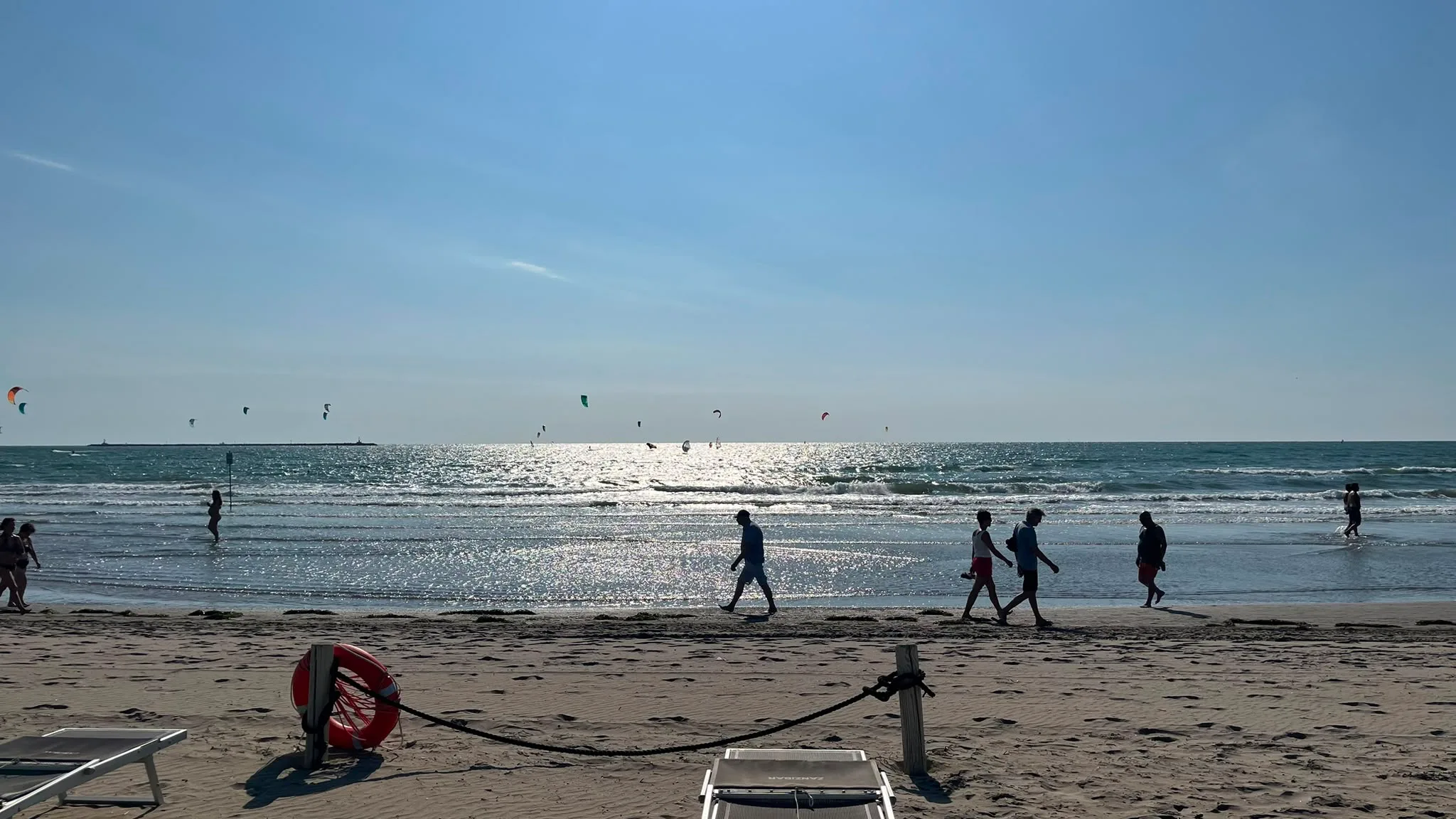 Panorama di spiaggia con lettini, ombrelloni, mare e numerosi kite surfisti all'orizzonte sotto cielo azzurro